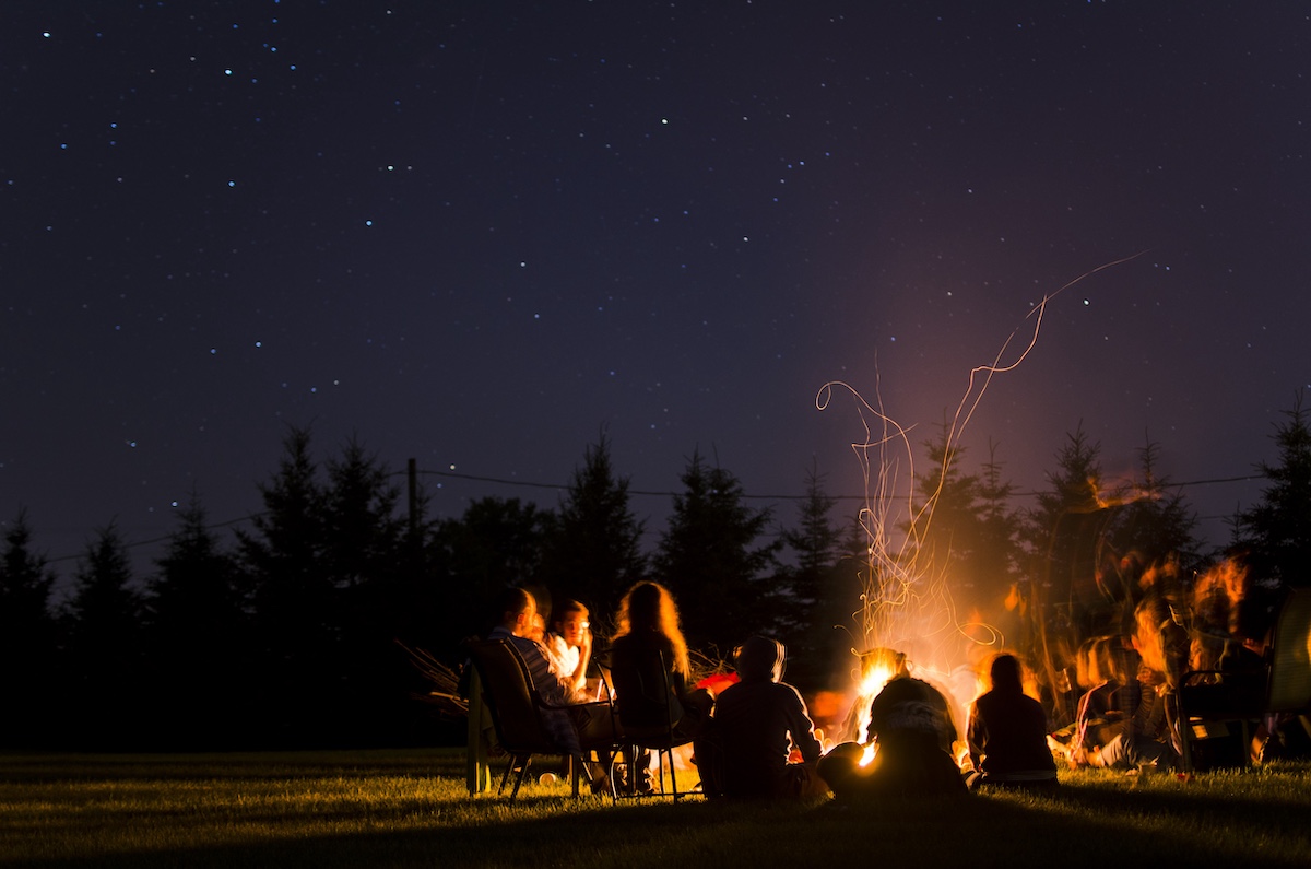 People sitting around a bonfire at night, evergreen trees in the distance